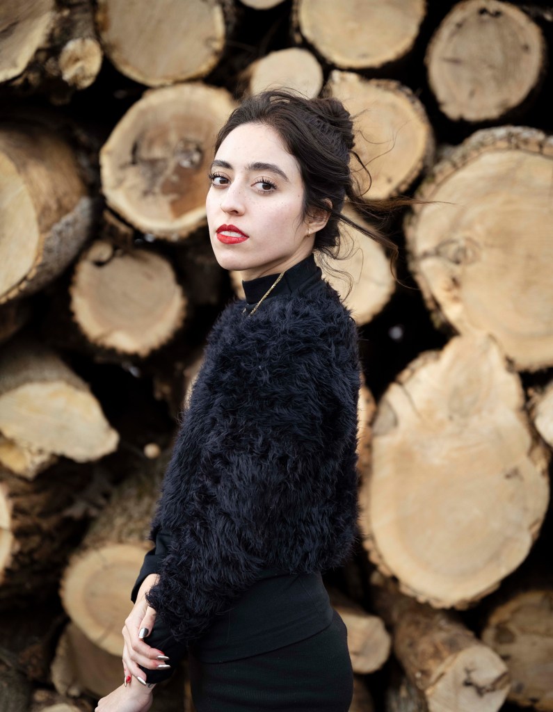 Portrait of a woman in black standing against stacked wooden logs.