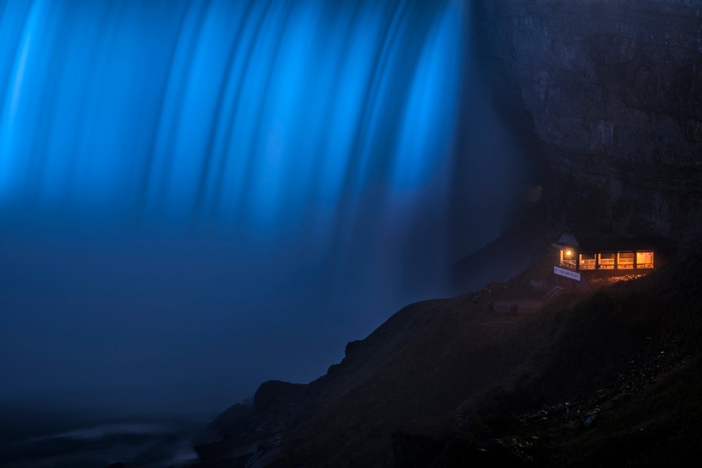 Niagara falls flowing through a blue-toned landscape with a small illuminated building nearby.