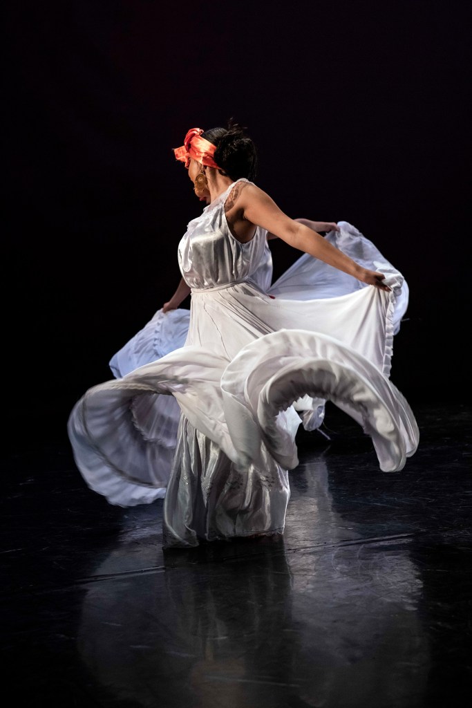 Dancer in a flowing white dress performing on a dark stage.